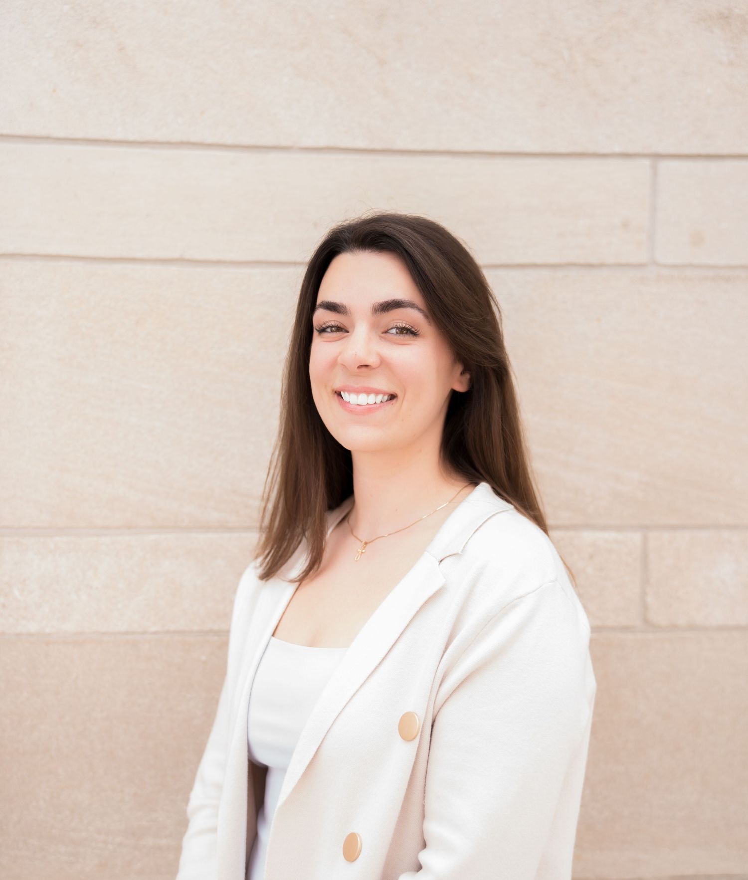 Young woman with long dark hair smiling in a white blazer, posing outdoors against a beige stone wall.
