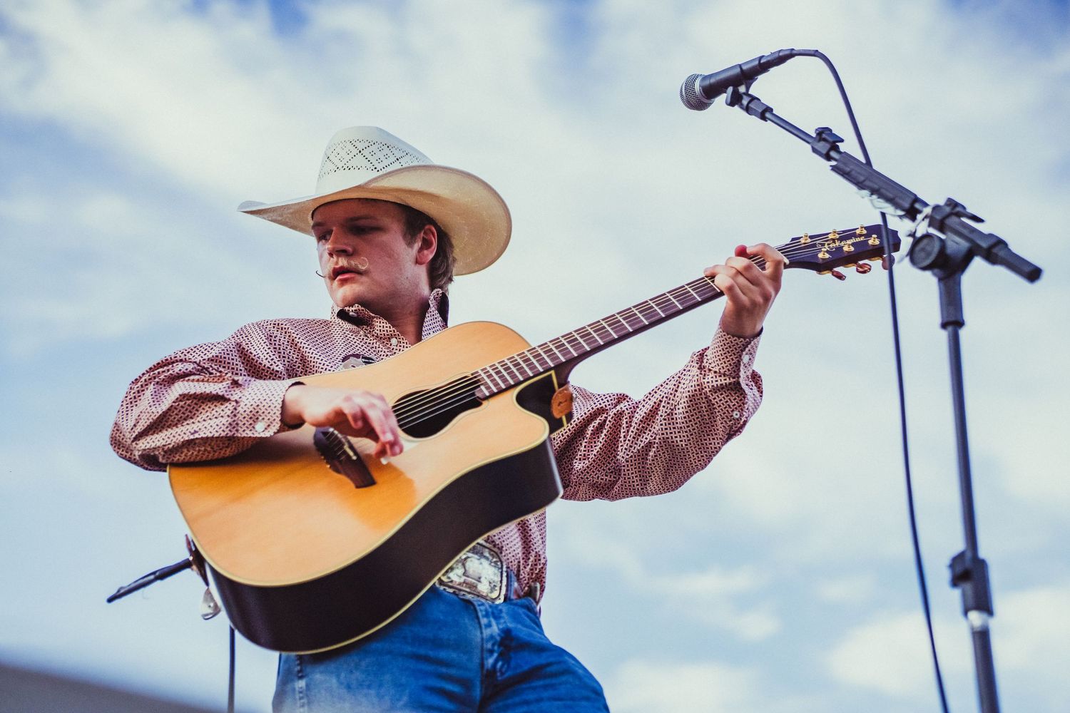 A young country musician in a white cowboy hat plays acoustic guitar on an outdoor stage.