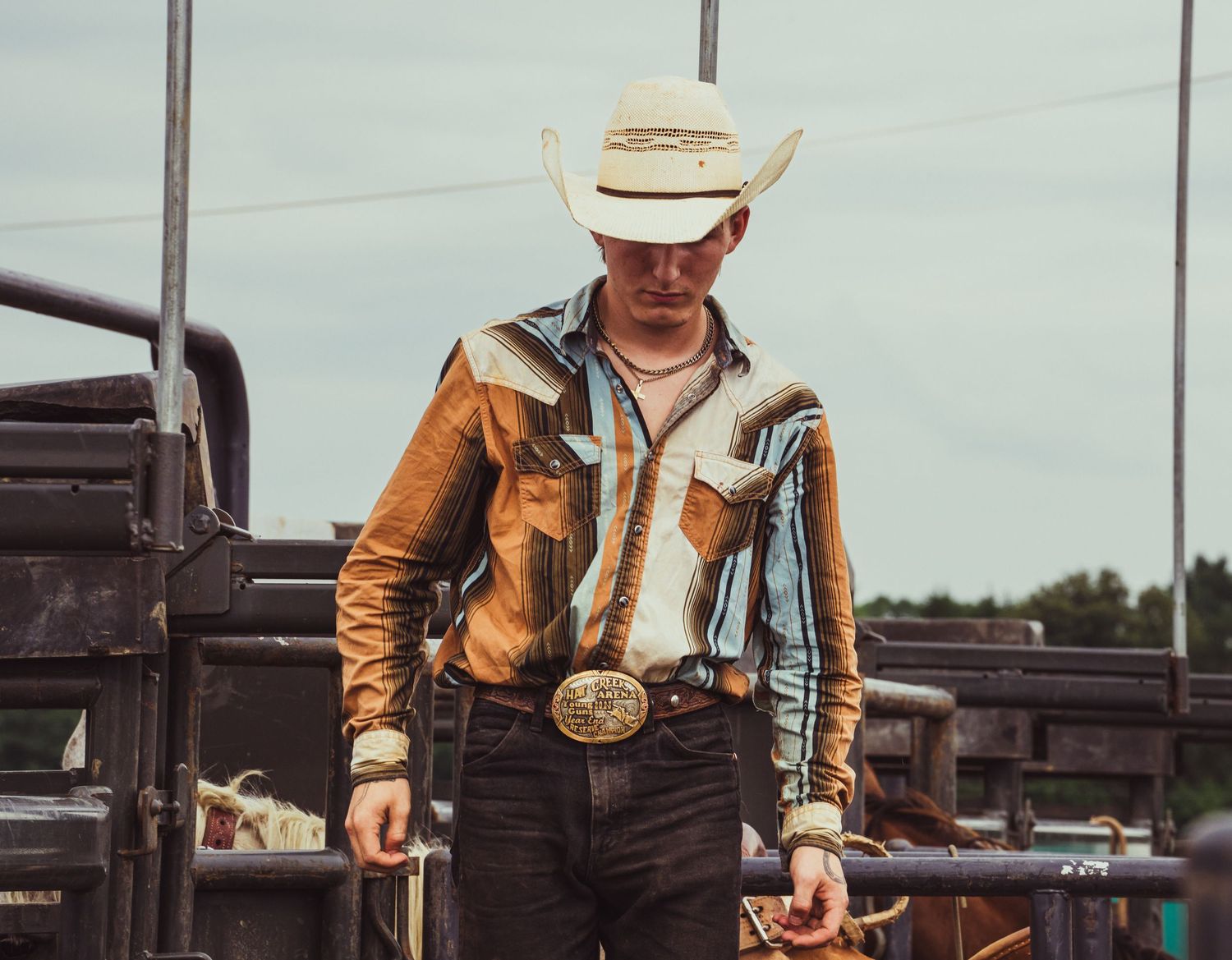 A cowboy in a white hat and Western shirt with a large belt buckle stands at a rodeo arena.