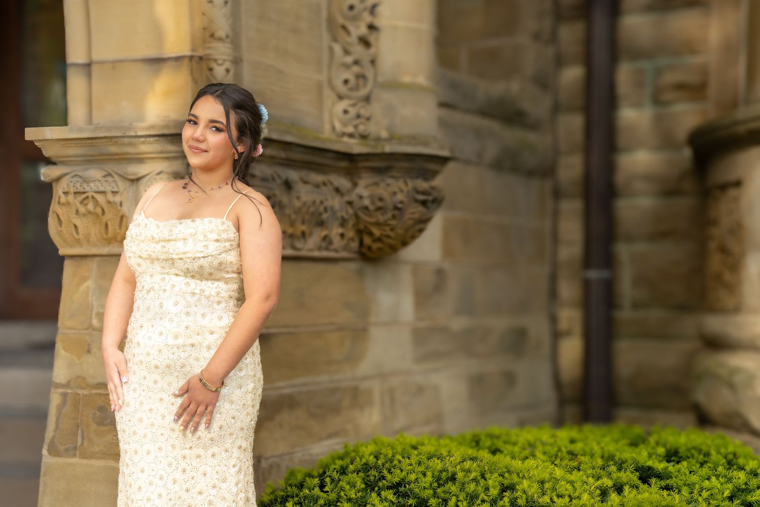 Elegant woman in a floral gown posing by ornate stone columns with lush greenery in the background.