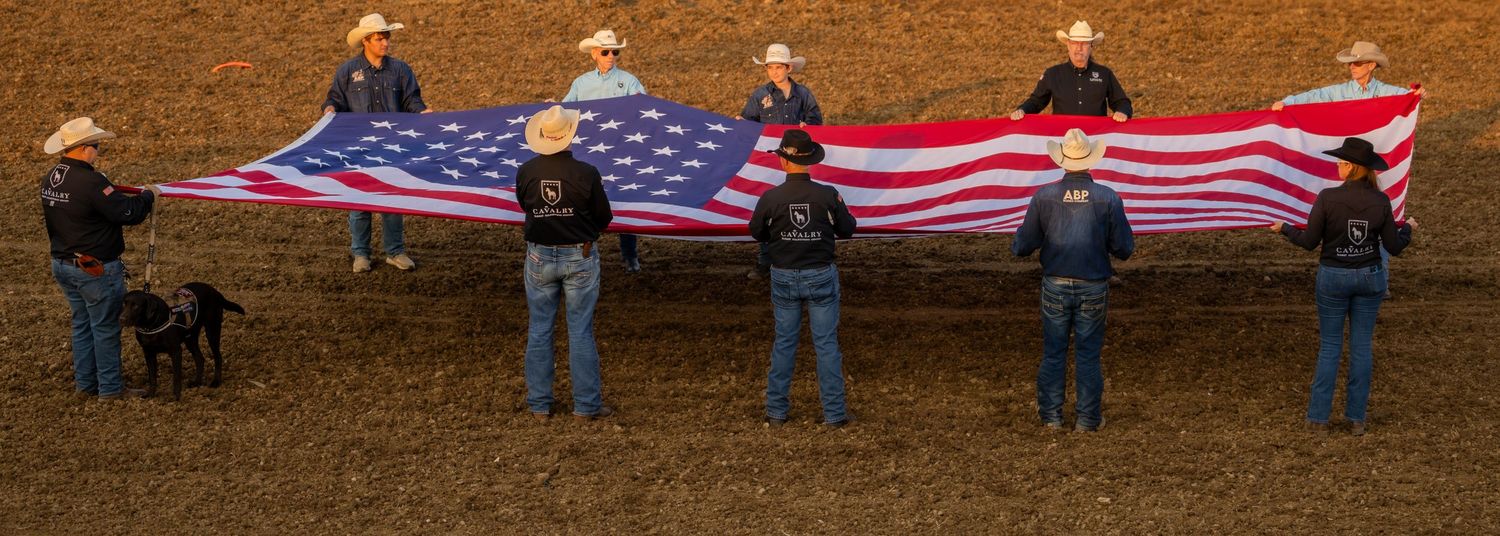 Group of people in cowboy hats holding a large American flag at an outdoor rodeo arena.