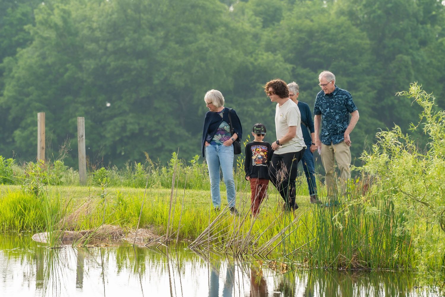 Family group of four adults and a child exploring a scenic pond shoreline surrounded by lush green vegetation.