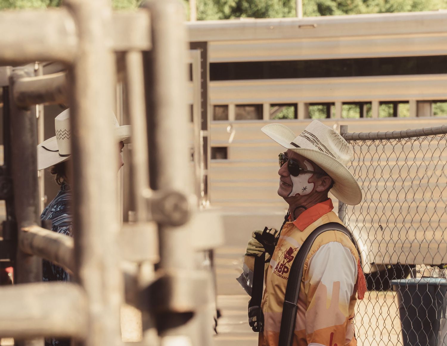 Rodeo clown in face paint and safety vest standing near chutes at outdoor rodeo.