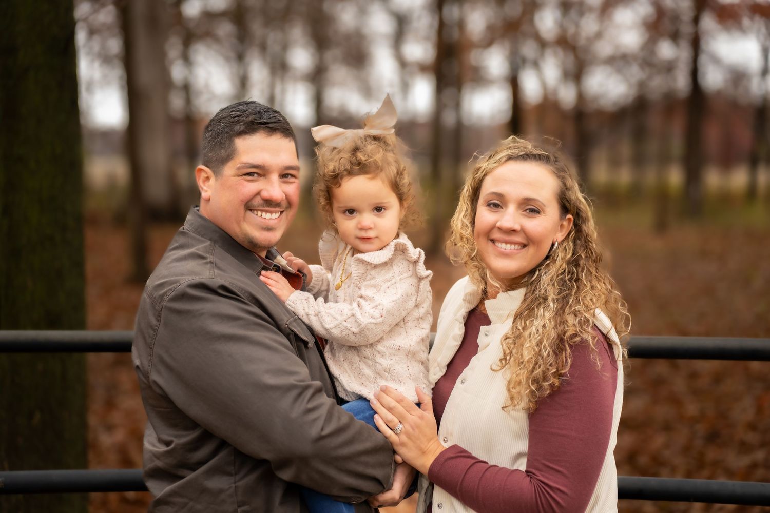 A smiling family of three poses outdoors in autumn, with a man holding a toddler girl beside a woman with curly hair.