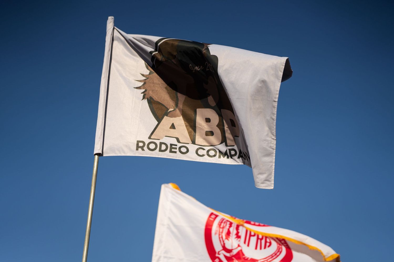ABE Rodeo Company branded white flag waves against a bright blue sky alongside a red championship flag.