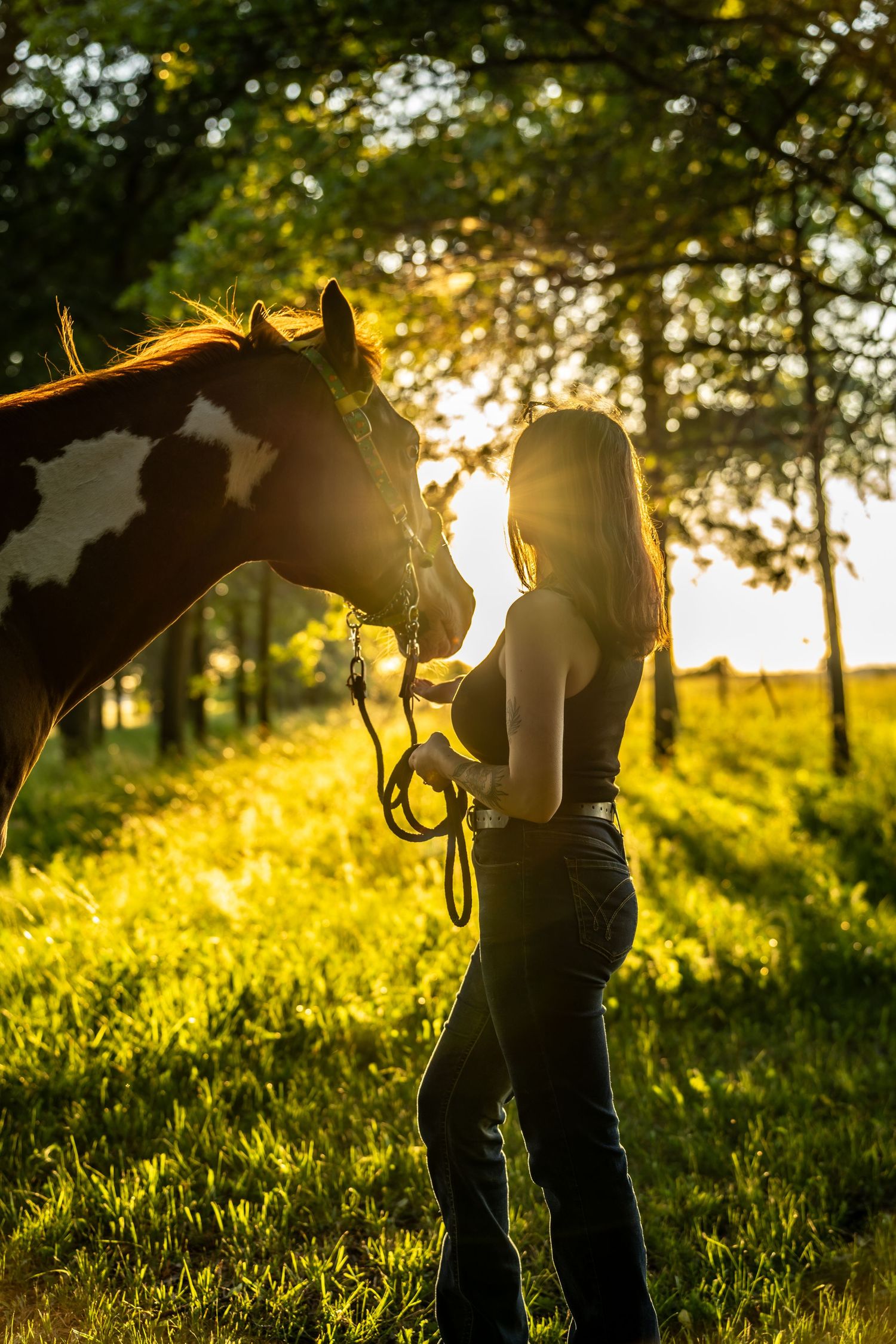 Woman standing with a paint horse in golden sunset light among green trees.