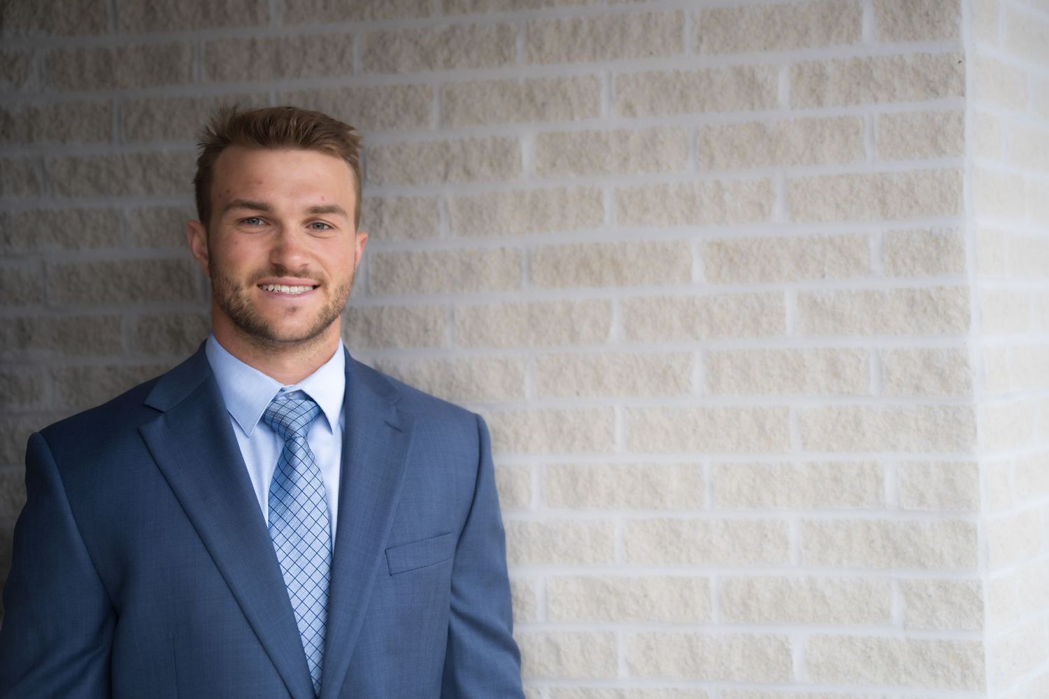 Professional young man in blue suit and tie smiling confidently against a light brick wall background.