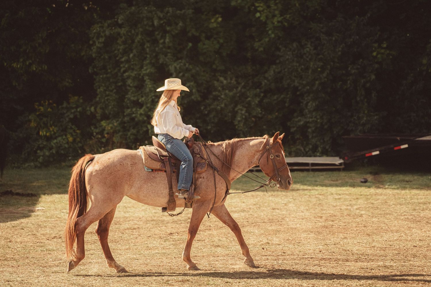 Blonde woman in cowboy hat riding chestnut horse in sunny outdoor ranch arena.
