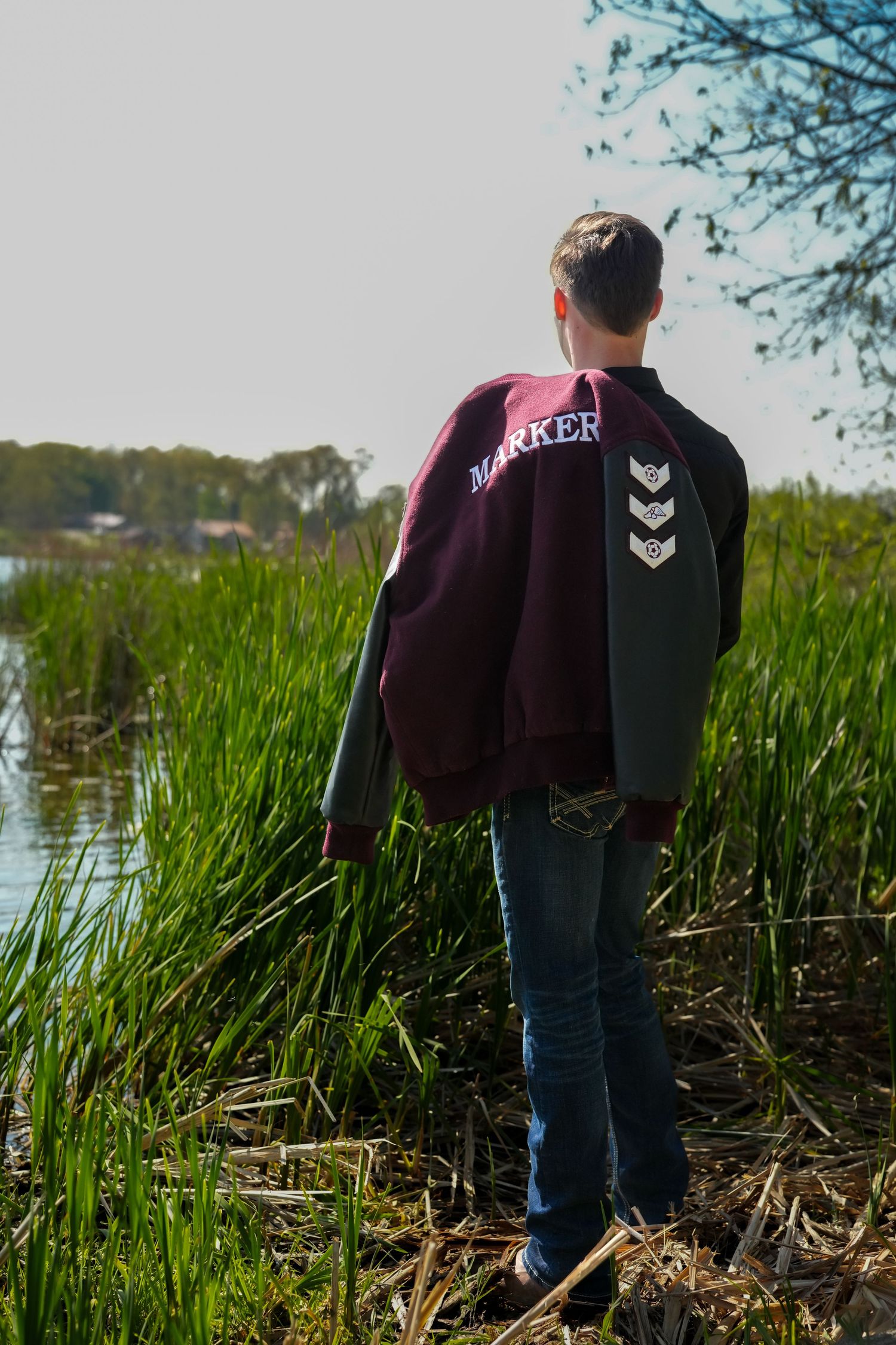 Young man wearing a Marker letterman jacket stands with his back to camera near a pond.