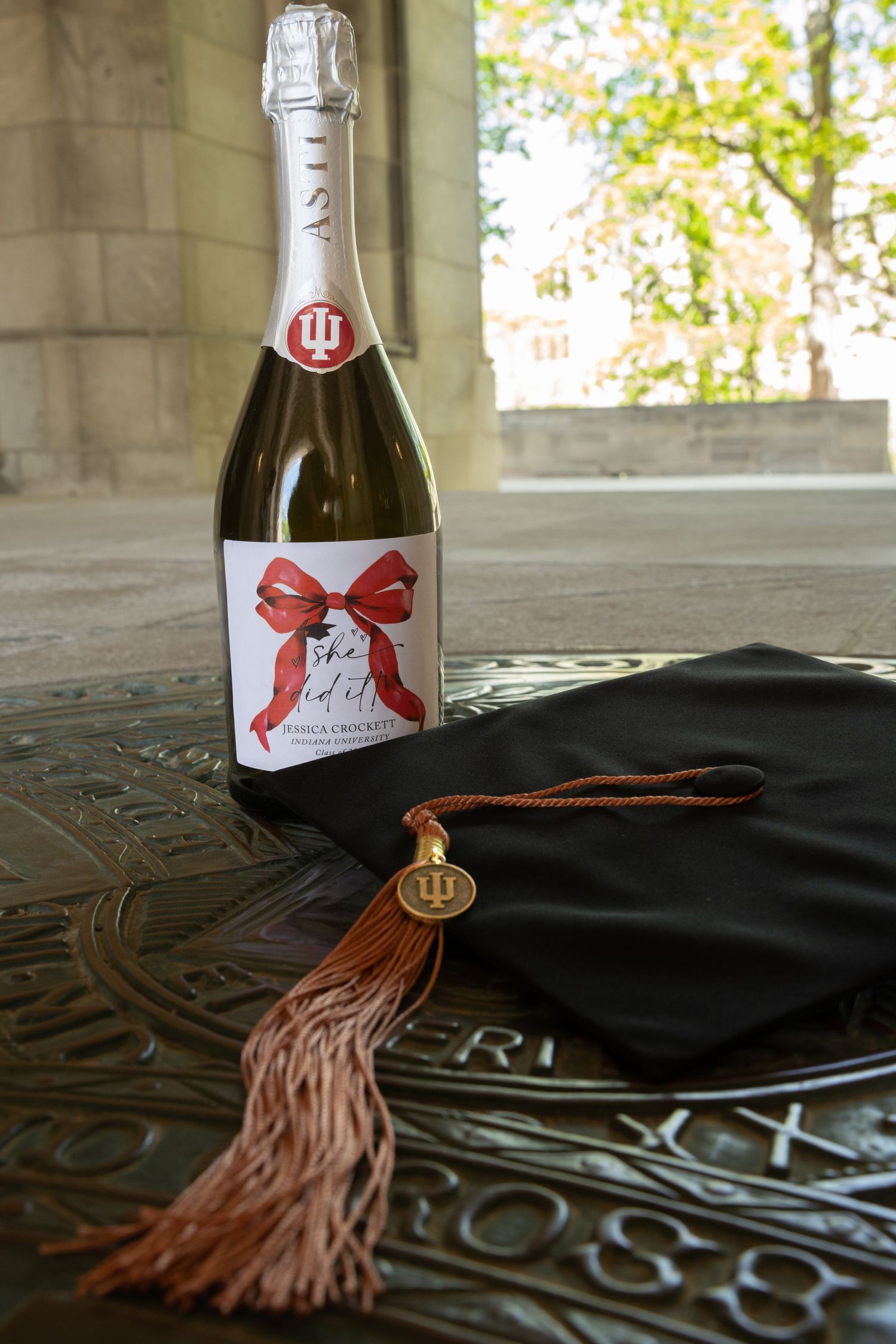 Indiana University graduation cap with IU tassel beside a custom sparkling wine bottle with red bow label on stone surface.