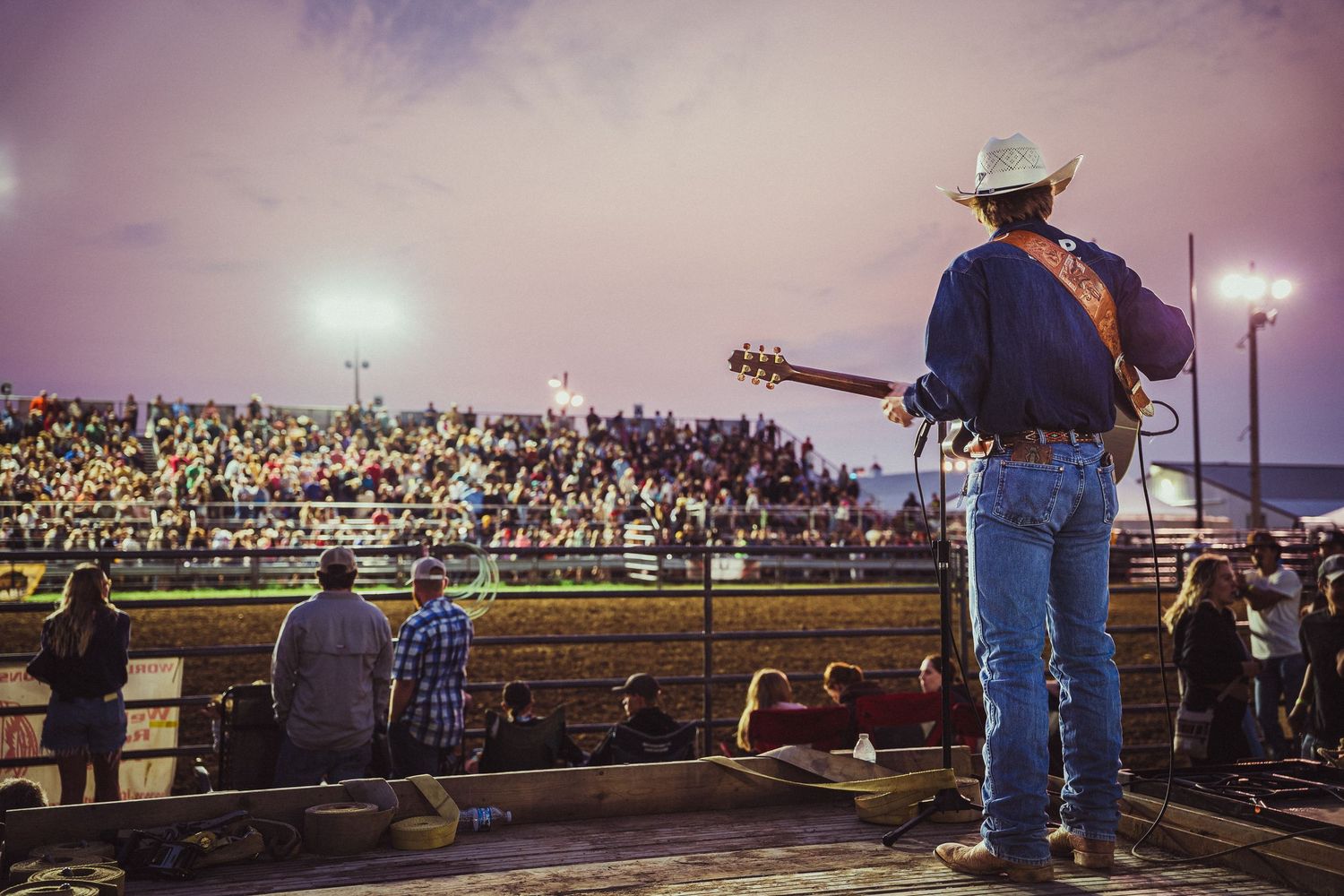 Country musician in cowboy hat plays guitar on stage at a packed outdoor rodeo arena at dusk.
