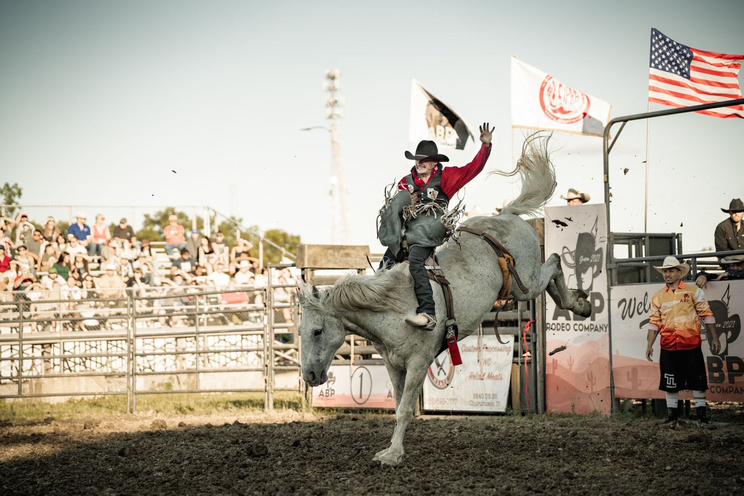Cowboy in black hat rides a bucking gray bronco near an American flag at a professional rodeo event.