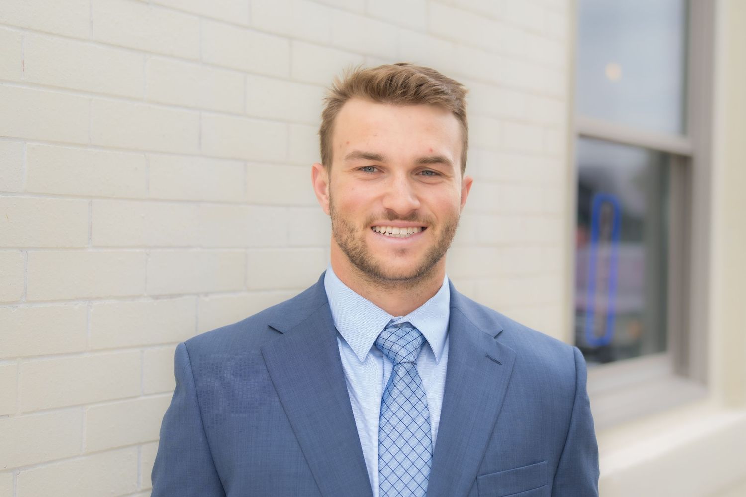 Young professional man in blue suit and tie smiling confidently against white brick wall outdoors.