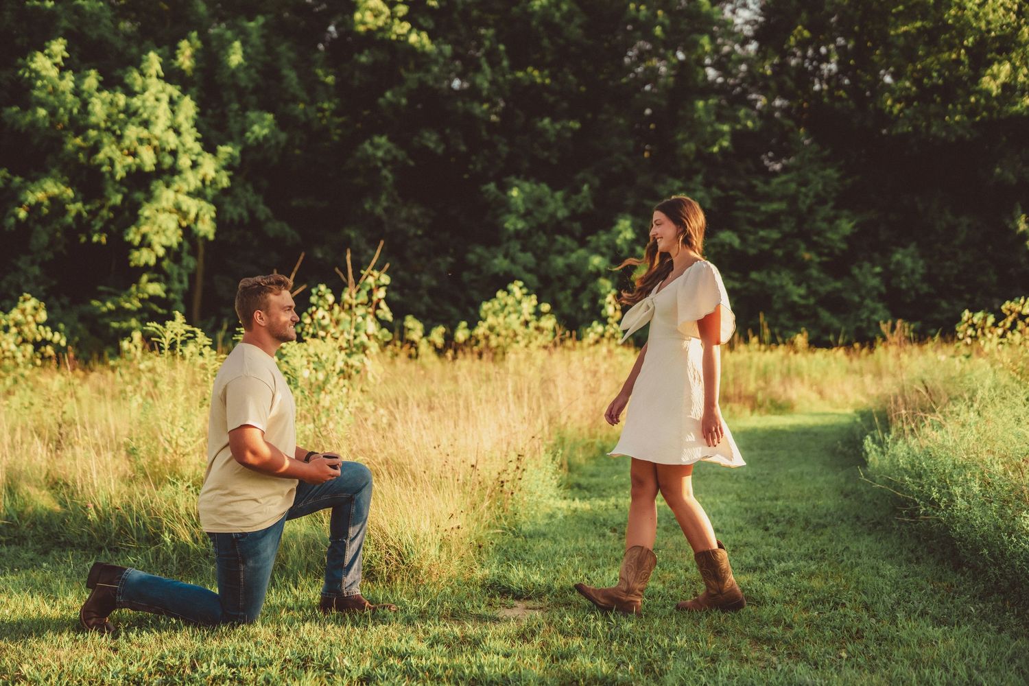 Man kneeling on one knee proposing to woman in white dress on a sunny green meadow path surrounded by trees.
