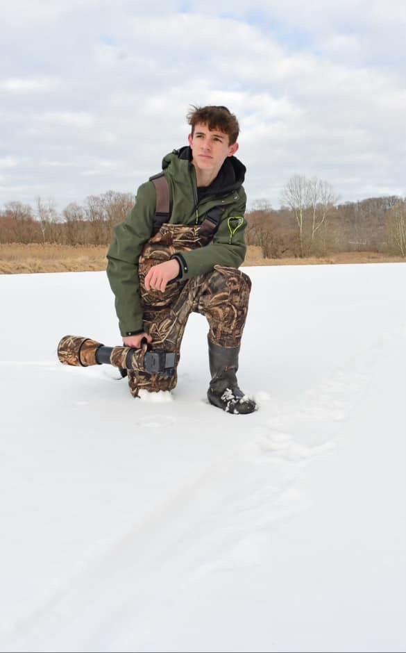 Young man in camo pants and green jacket kneeling on frozen lake holding camera lens in winter landscape.