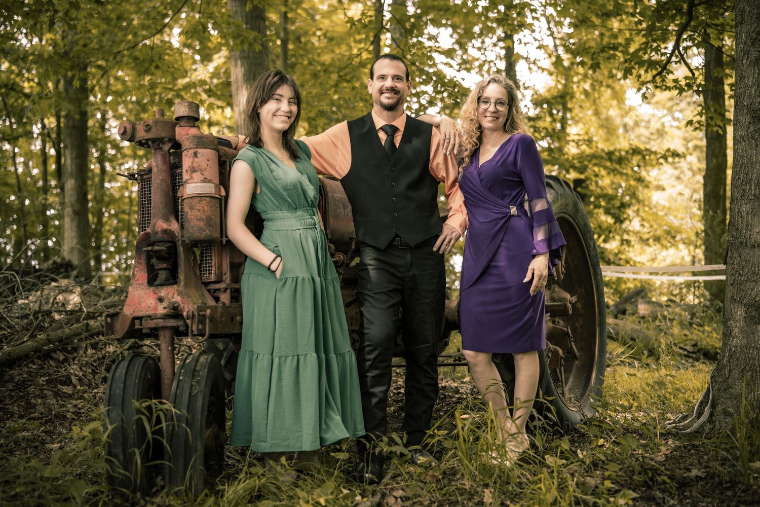 Three musicians pose outdoors near a vintage tractor surrounded by lush green trees, dressed in elegant green and purple attire.