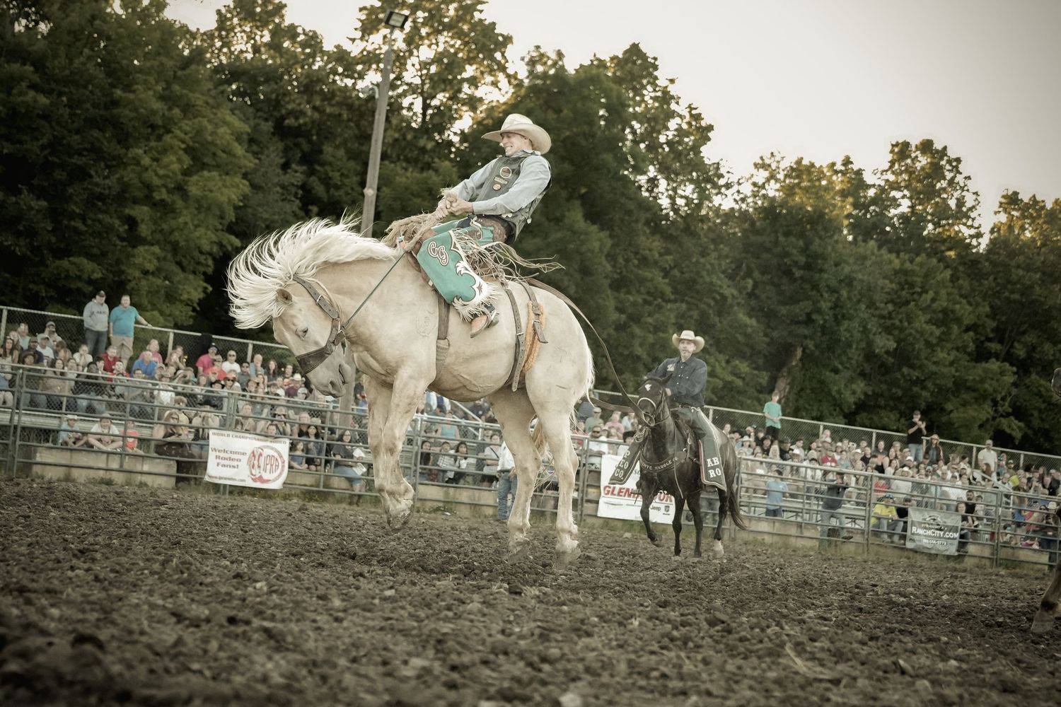 Rodeo rider on a white horse performing in a dirt arena with spectators in the stands.