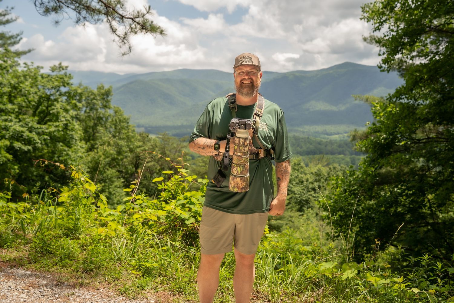 Man with camera and gear stands on mountain trail surrounded by lush greenery with scenic mountain range in background.