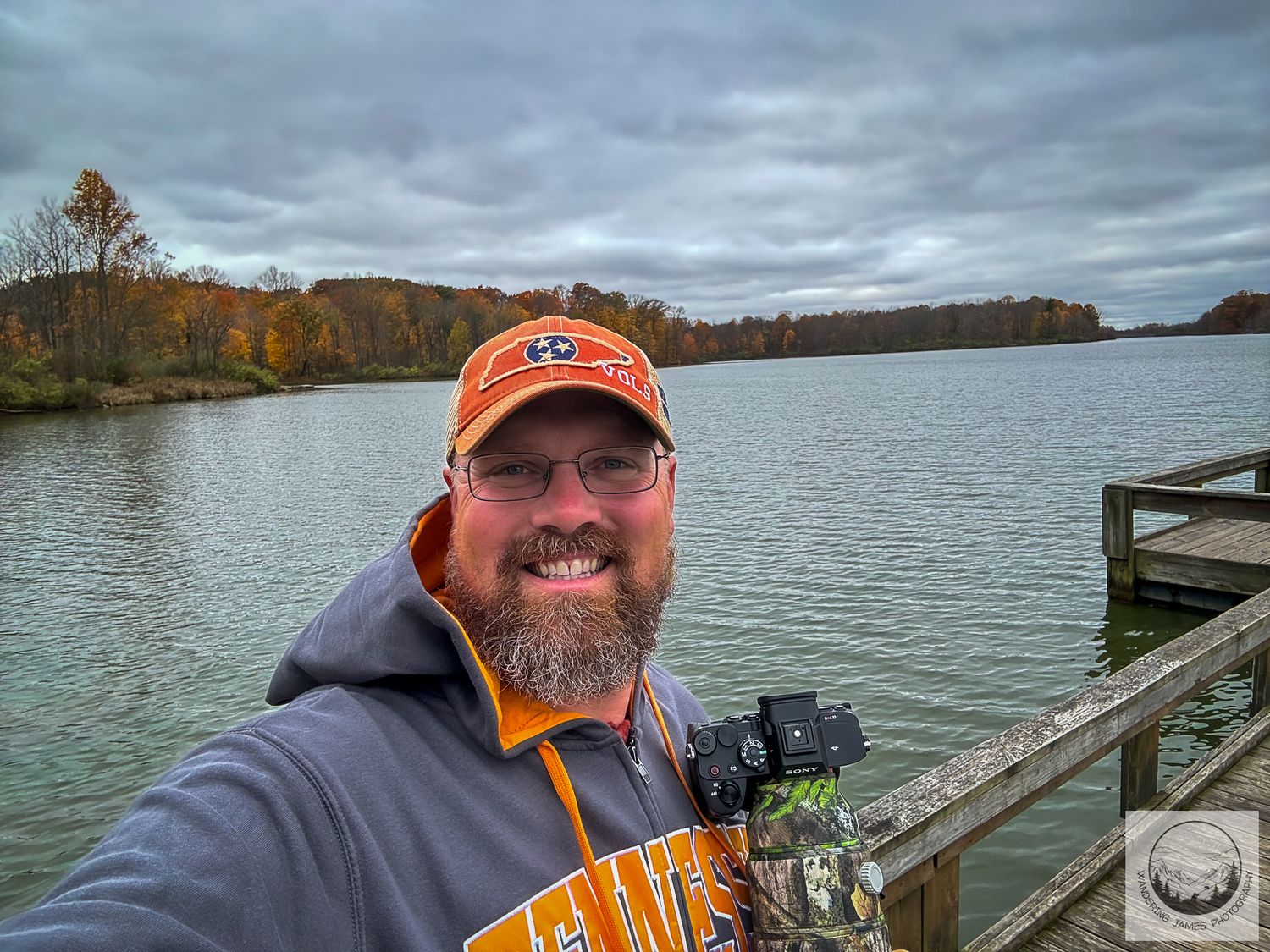 Man taking selfie on lake dock wearing orange cap and hoodie, with autumn trees and cloudy sky reflected on calm water.