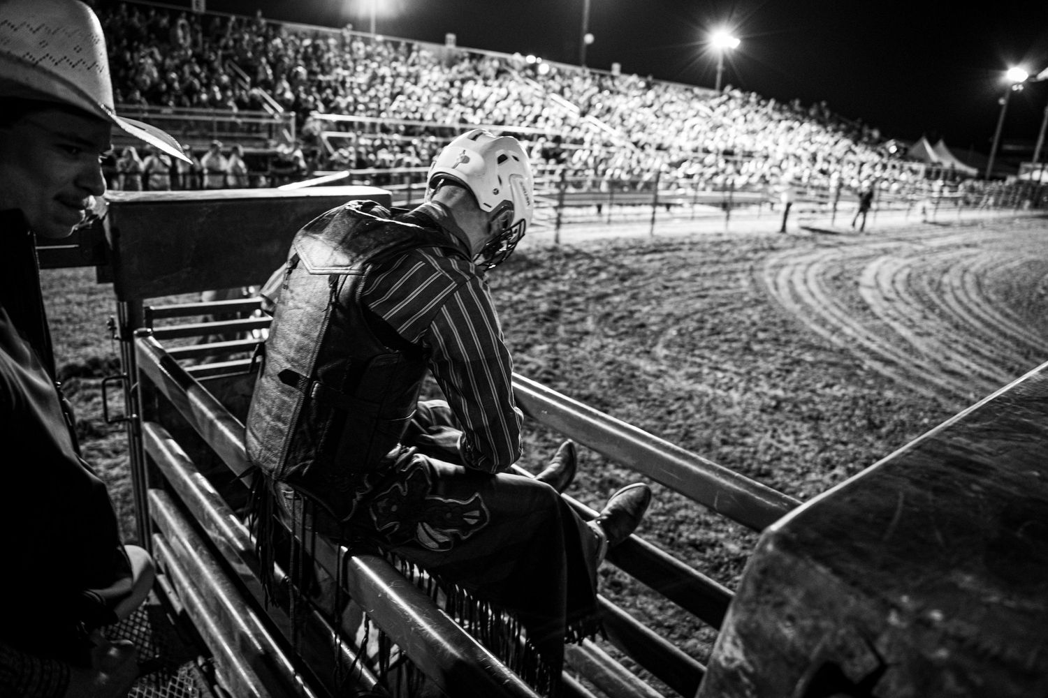 Black and white rodeo photo of a bull rider preparing in the chute at a packed night arena.