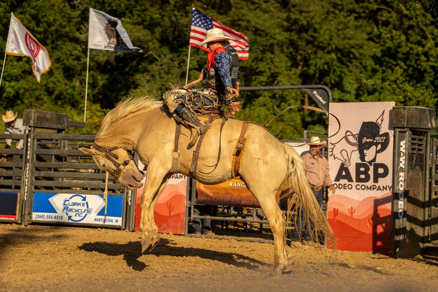 A cowboy rides a palomino bucking horse near an American flag at an ABP Rodeo Company event.