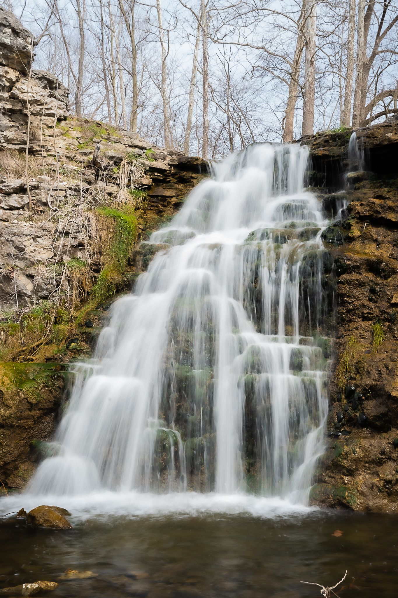 Cascading waterfall flowing over rocky ledge surrounded by bare trees in spring.
