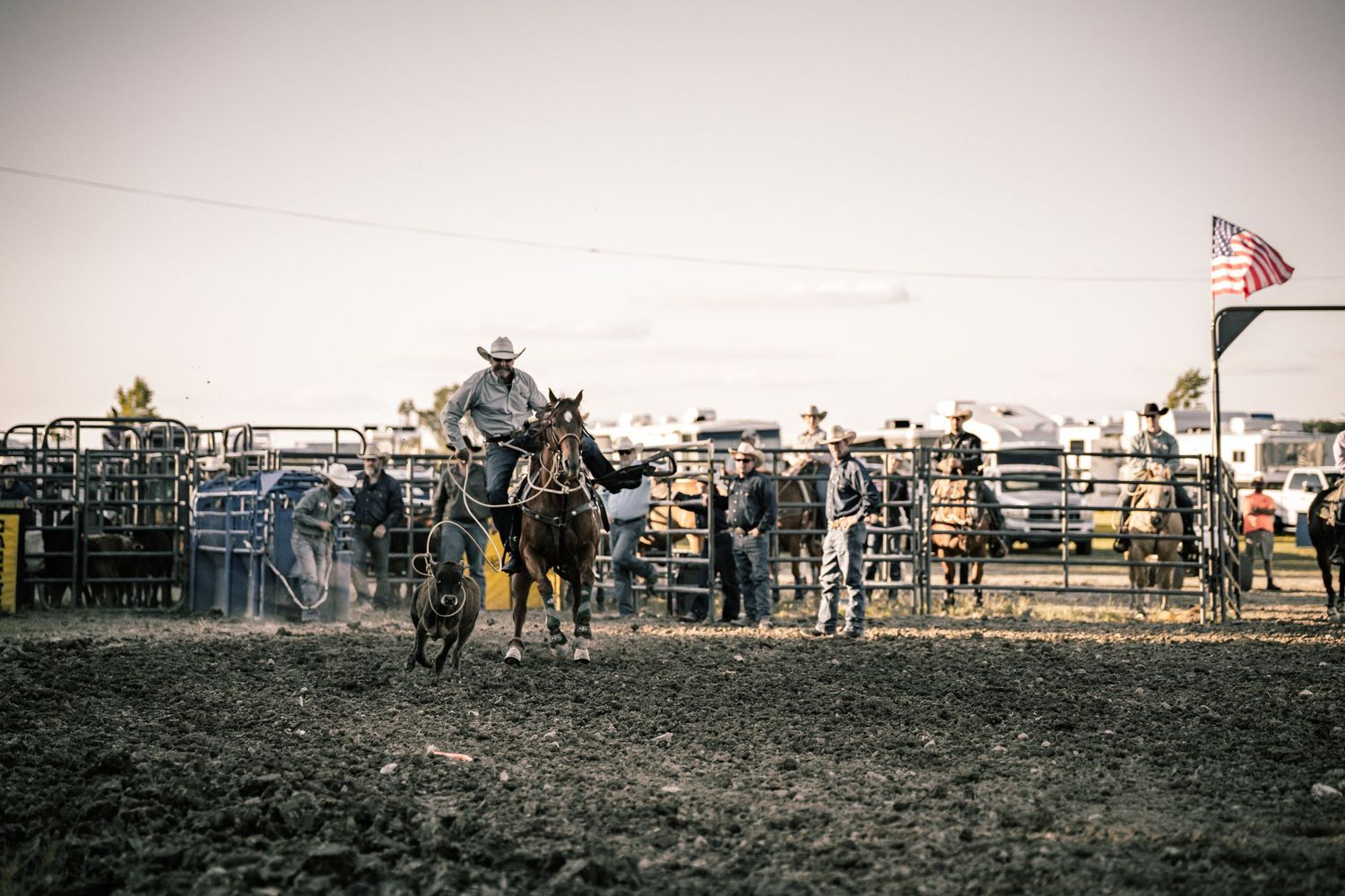Cowboy on horseback in a rodeo arena at dusk with an American flag and spectators in the background.