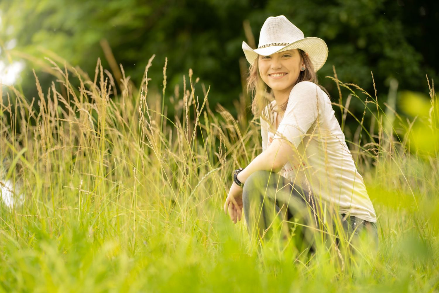 Smiling young woman in a white cowboy hat sitting in tall green grass on a sunny day.