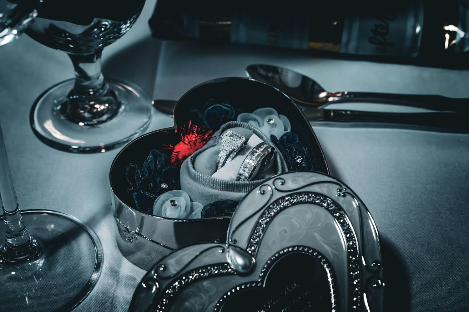 Heart-shaped silver jewelry box with skull and red rose decorations, surrounded by wine glasses on a dark surface.