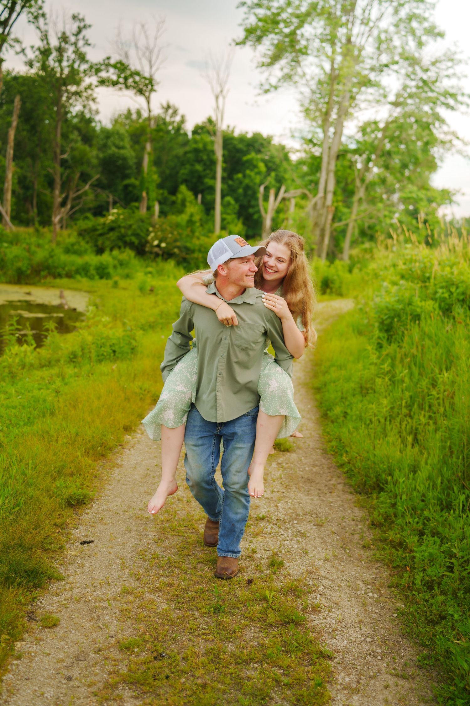 A young couple laughing on a rural path, man giving woman a piggyback ride surrounded by lush green trees.