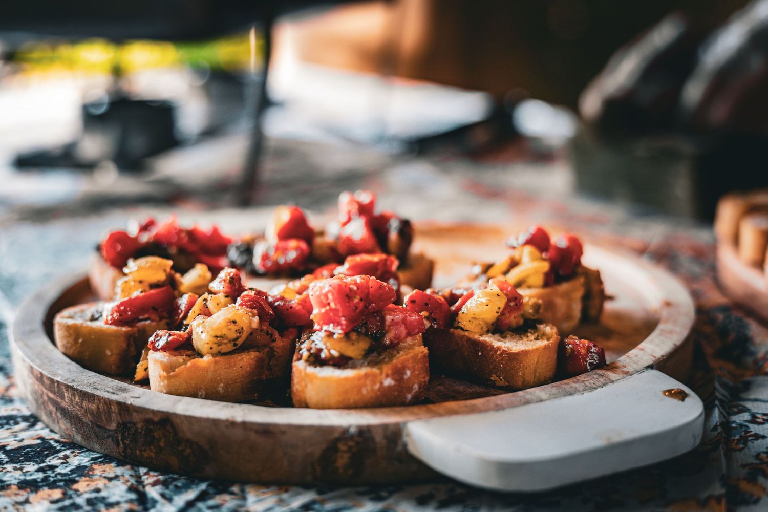 Rustic wooden platter of bruschetta topped with tomatoes and roasted vegetables on a decorative table.