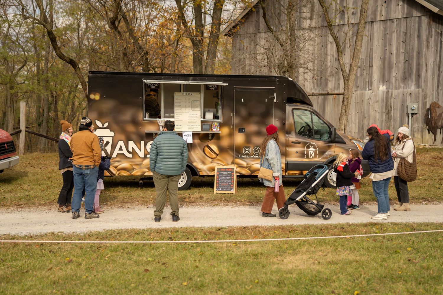 Customers line up at a dark food truck with bread graphics in an outdoor autumn setting near a barn.