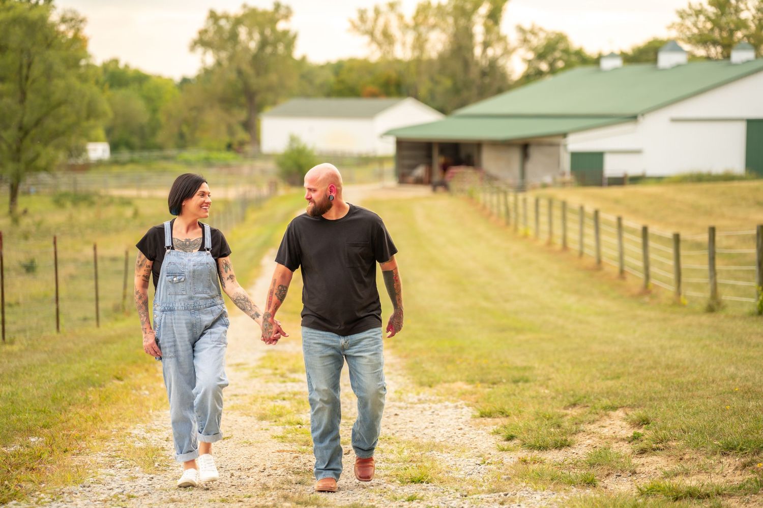 Couple holding hands walking down a gravel farm path with a barn in the background.