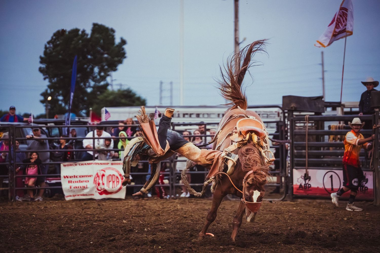 A rodeo cowboy rides a bucking bronco at an outdoor rodeo event with spectators watching in the background.
