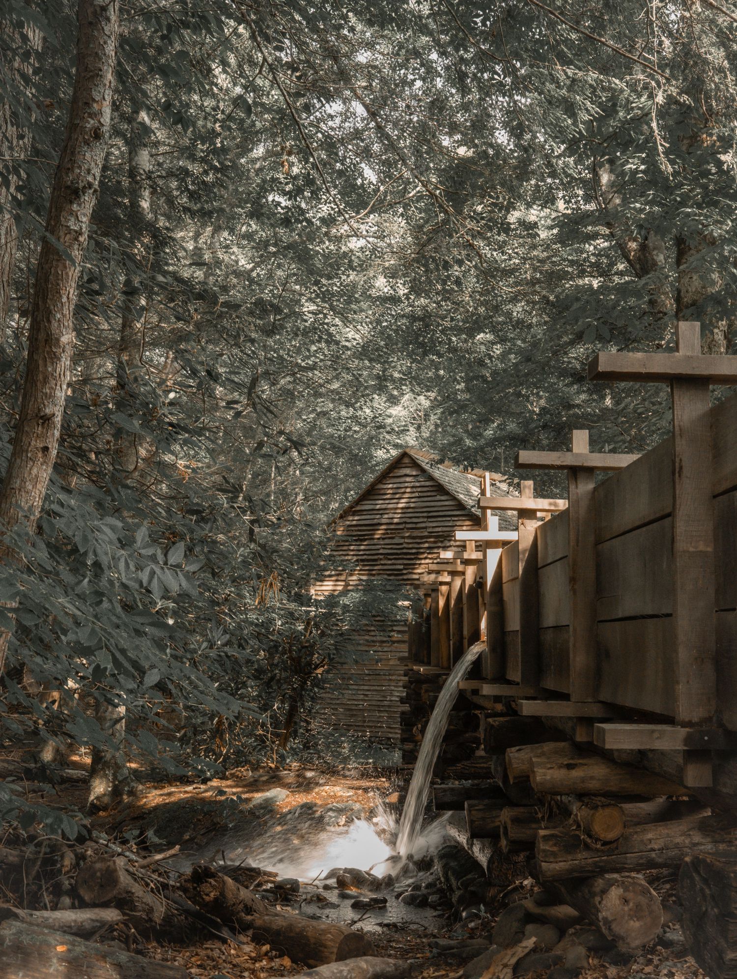 A rustic wooden grist mill with a water wheel surrounded by lush green trees in a serene forest setting.