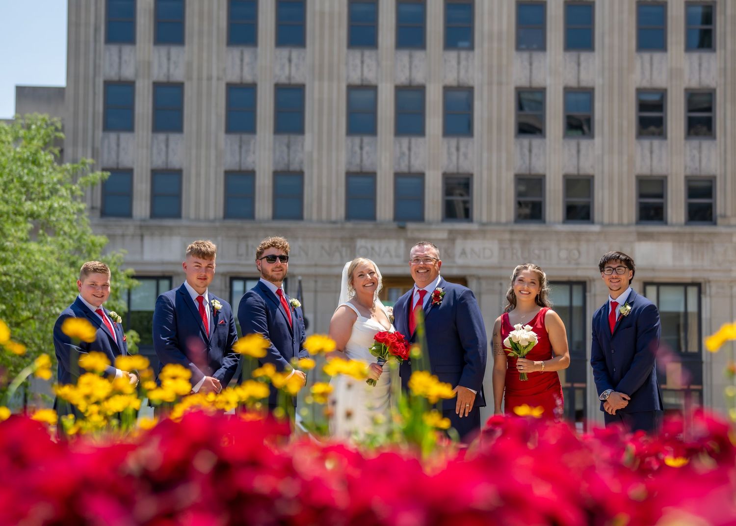 Wedding party poses outdoors with red and yellow flowers, groomsmen in navy suits with red ties flanking bride and groom.