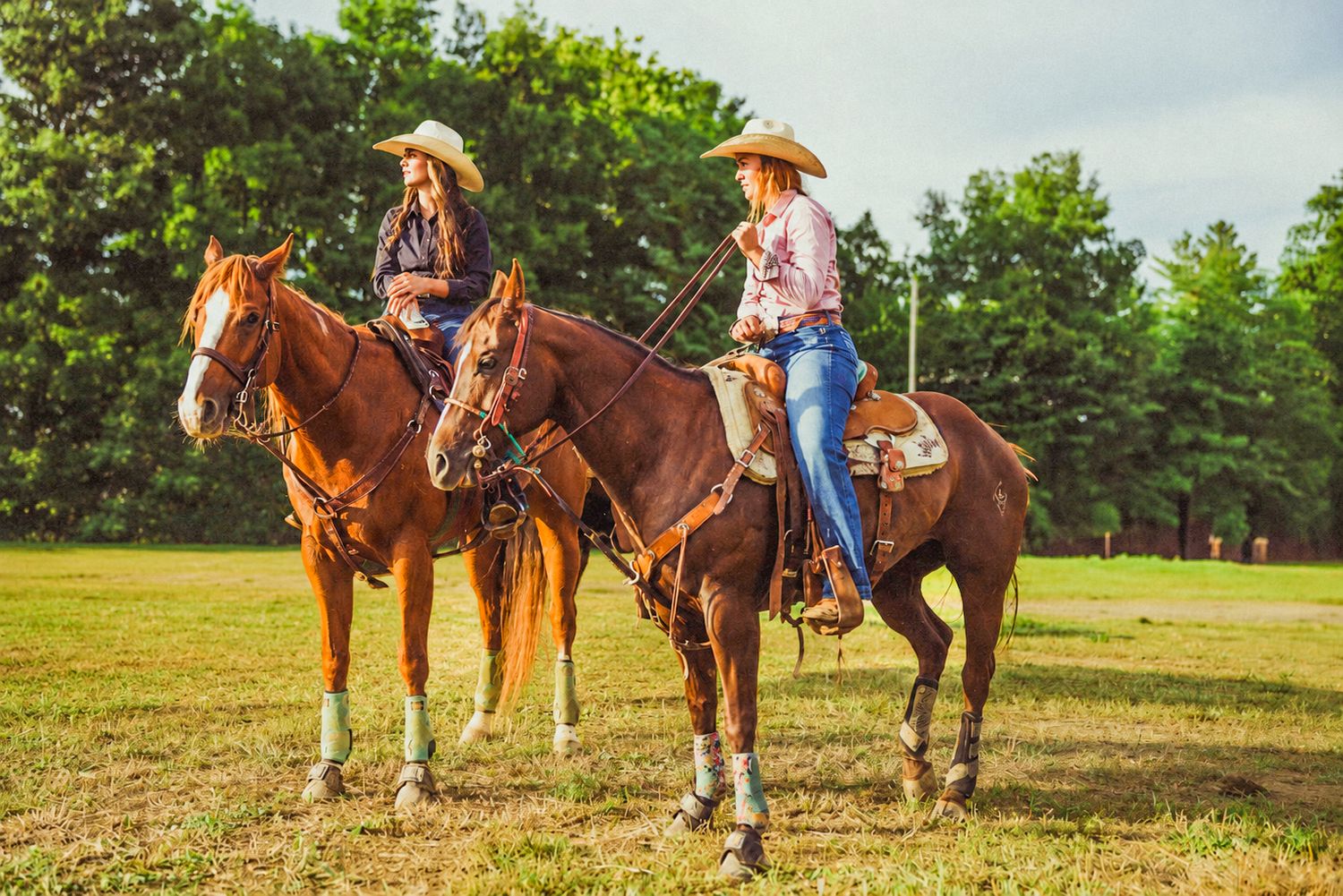 Two cowgirls in hats ride chestnut horses on a green field with trees in the background.