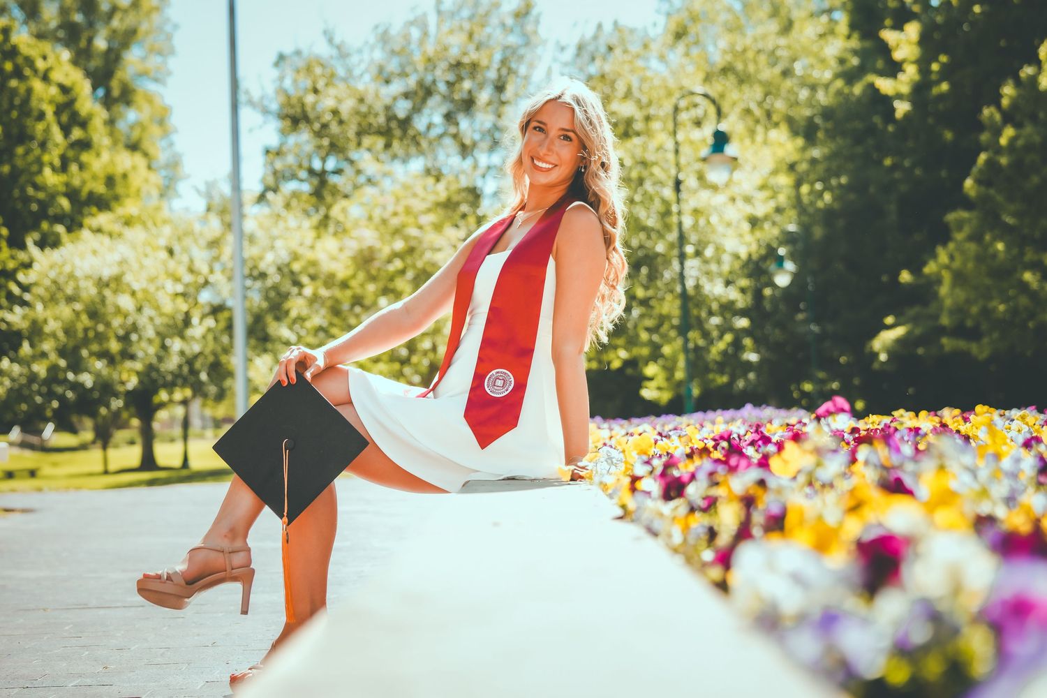 Smiling graduate in white dress and cap sitting beside colorful flowers outdoors.