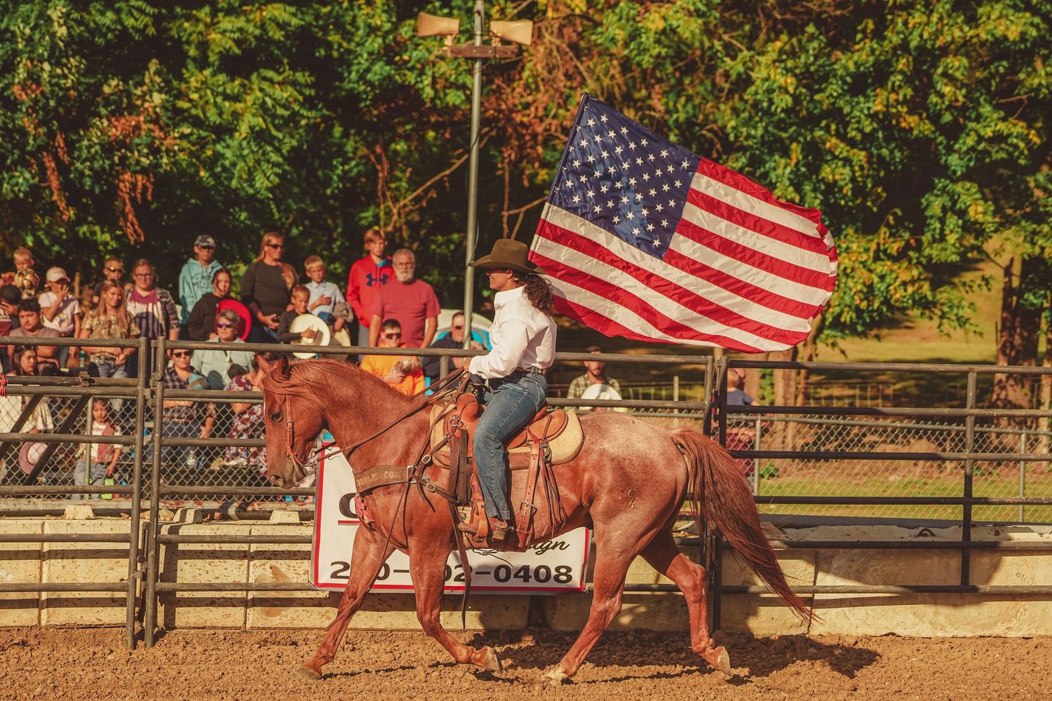 Rider on chestnut horse carries a large American flag past spectators at an outdoor rodeo arena.