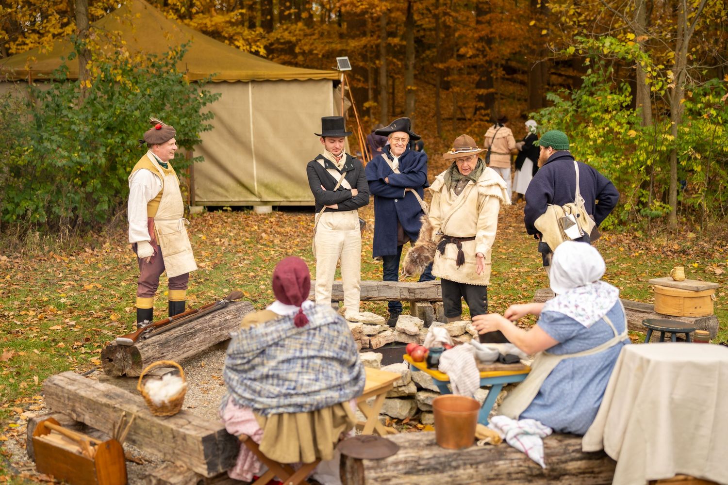 Historical reenactors in period costumes gather around a rustic outdoor camp with autumn foliage backdrop.