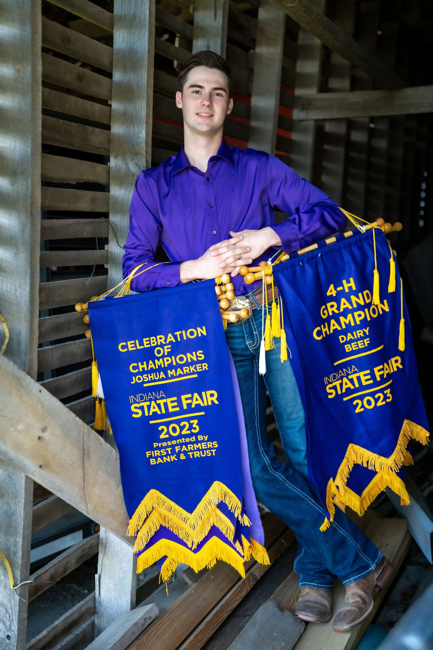 Young man in purple shirt holding 4-H Grand Champion Dairy Beef Indiana State Fair 2023 banners on stairs.