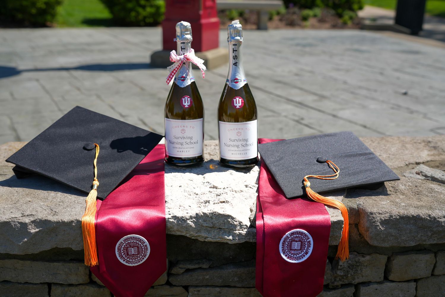 Two champagne bottles with graduation caps and crimson Indiana University stoles on stone surface.