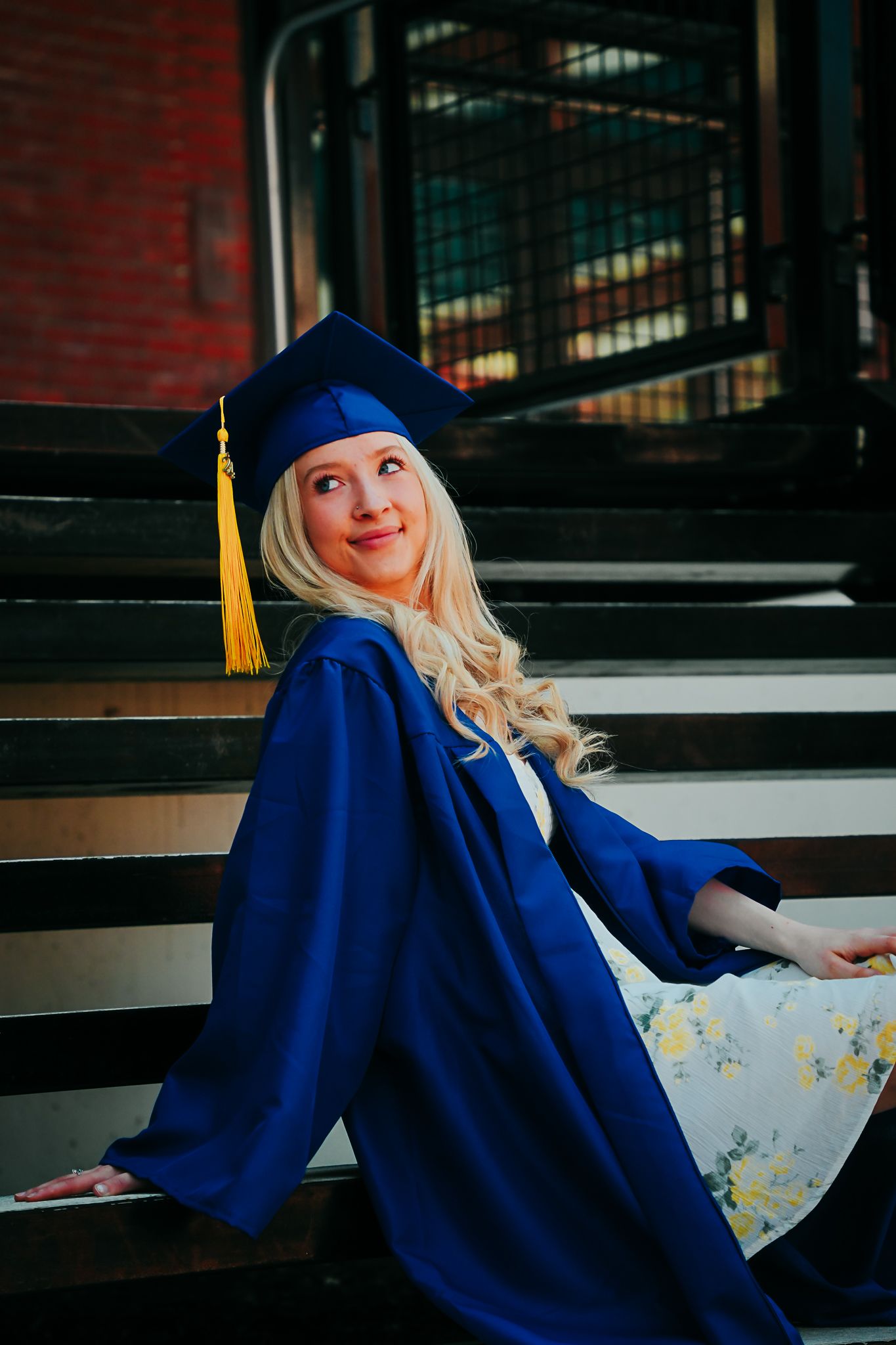 Blonde graduate in blue cap and gown sitting on steps looking up thoughtfully.
