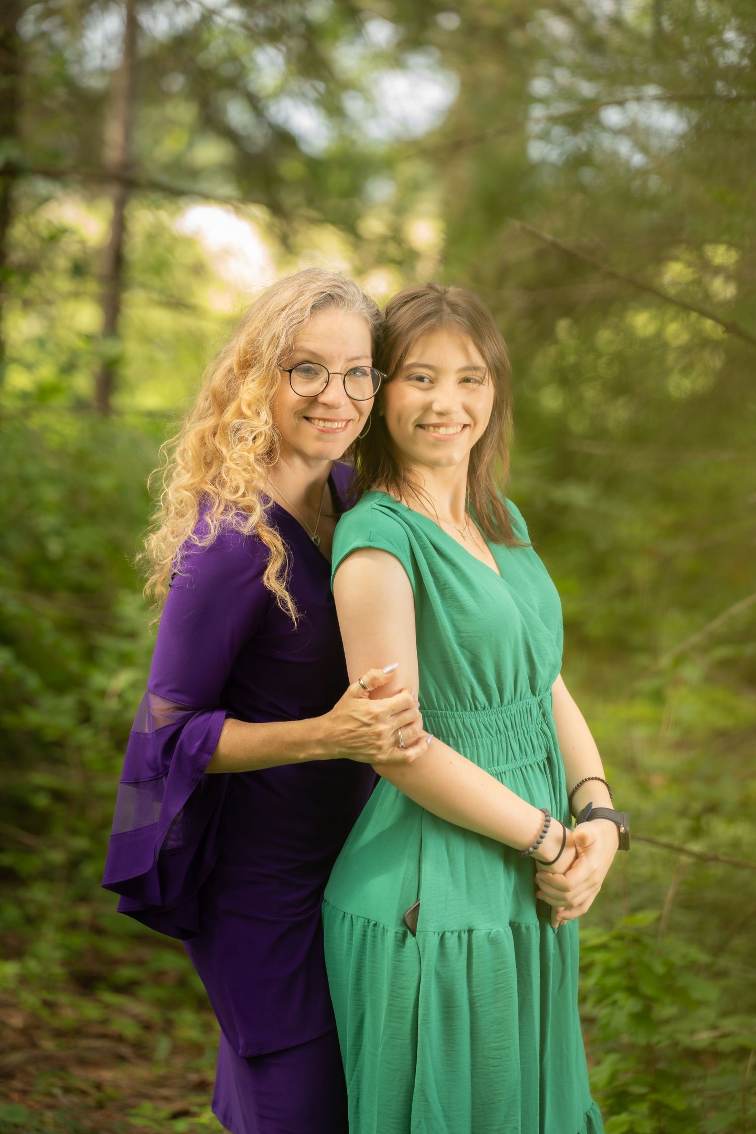 Two women in purple and green dresses pose together smiling in a lush green forest setting.