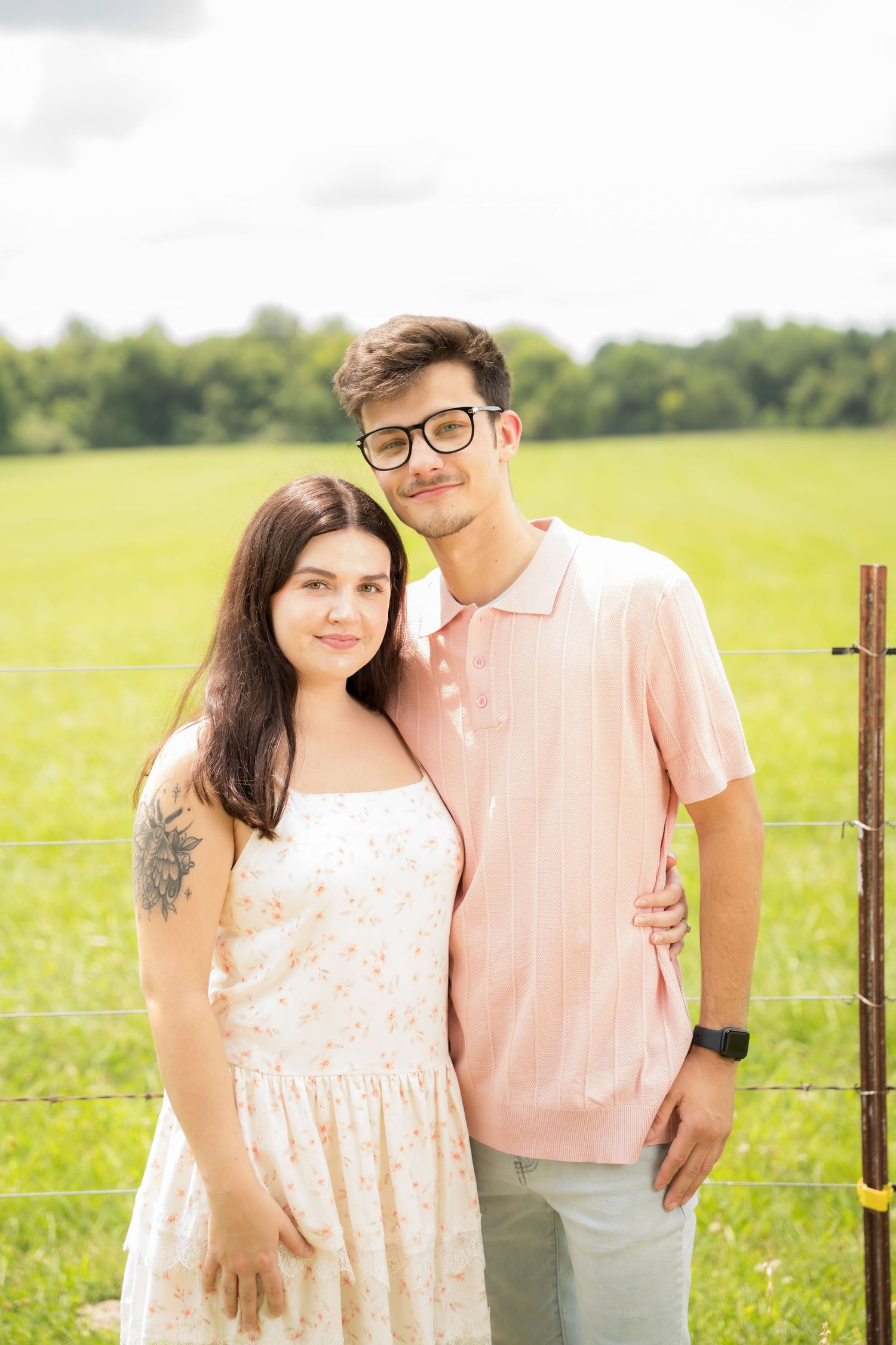 Young couple posing together in a green field, woman in floral dress with tattoo, man in pink polo with glasses.