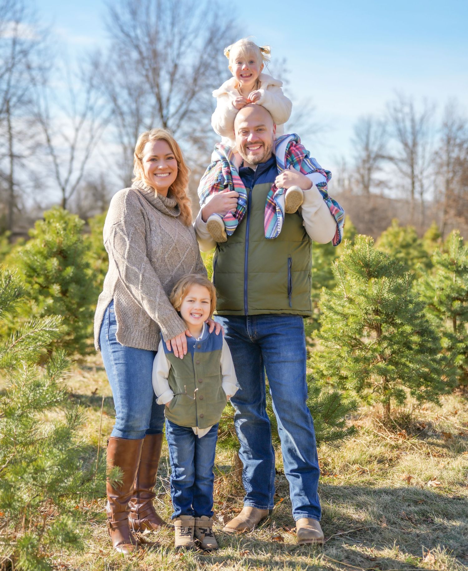 A happy family of four poses among Christmas trees, with dad carrying a toddler on his shoulders.