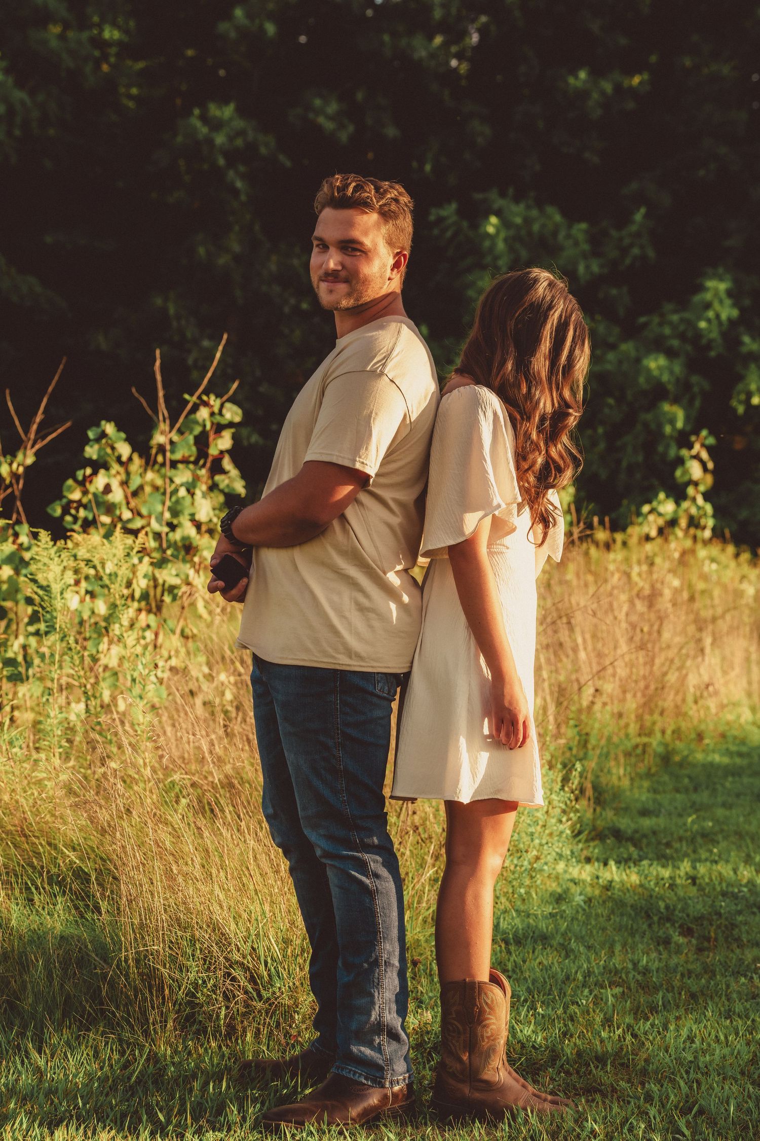 Couple standing back-to-back in a sunlit meadow, man smiling over shoulder, woman in white dress and cowboy boots.