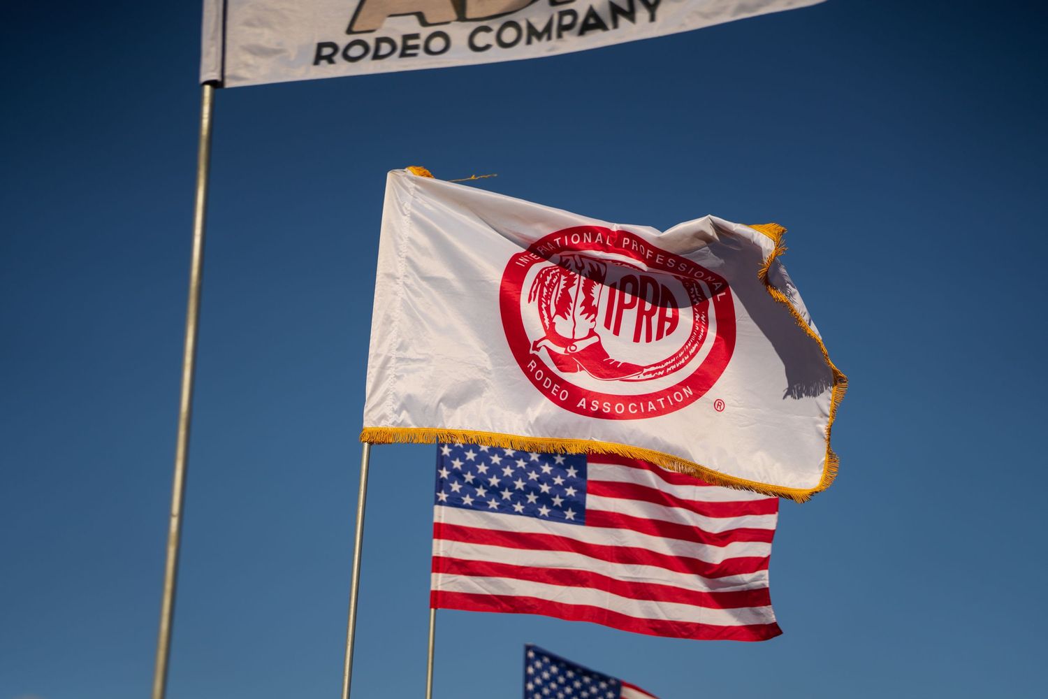 Rodeo Company and American flags flying against a clear blue sky at an outdoor rodeo event.
