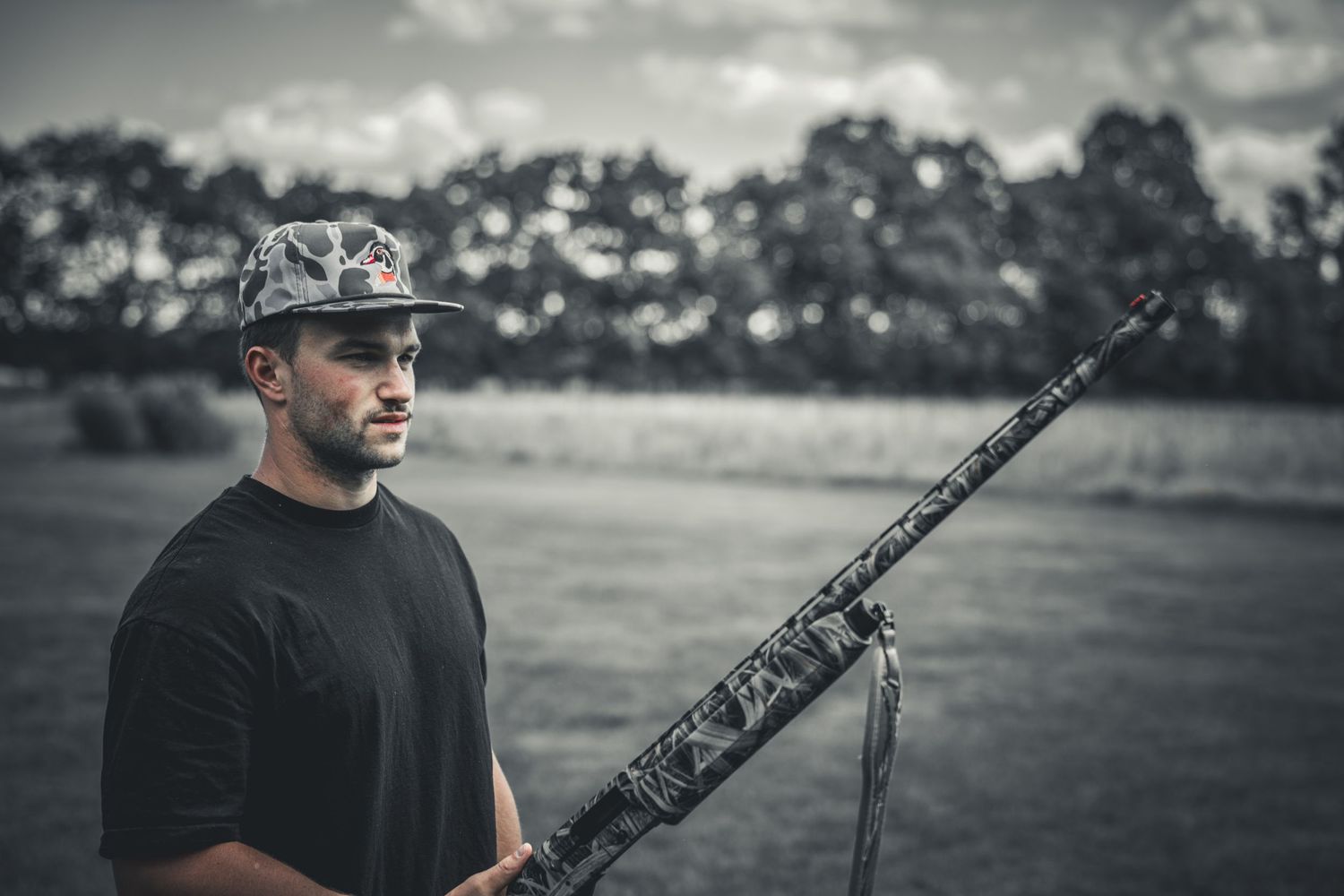 Man in camo hat holding a camouflage-patterned rifle outdoors in a field with trees in the background.