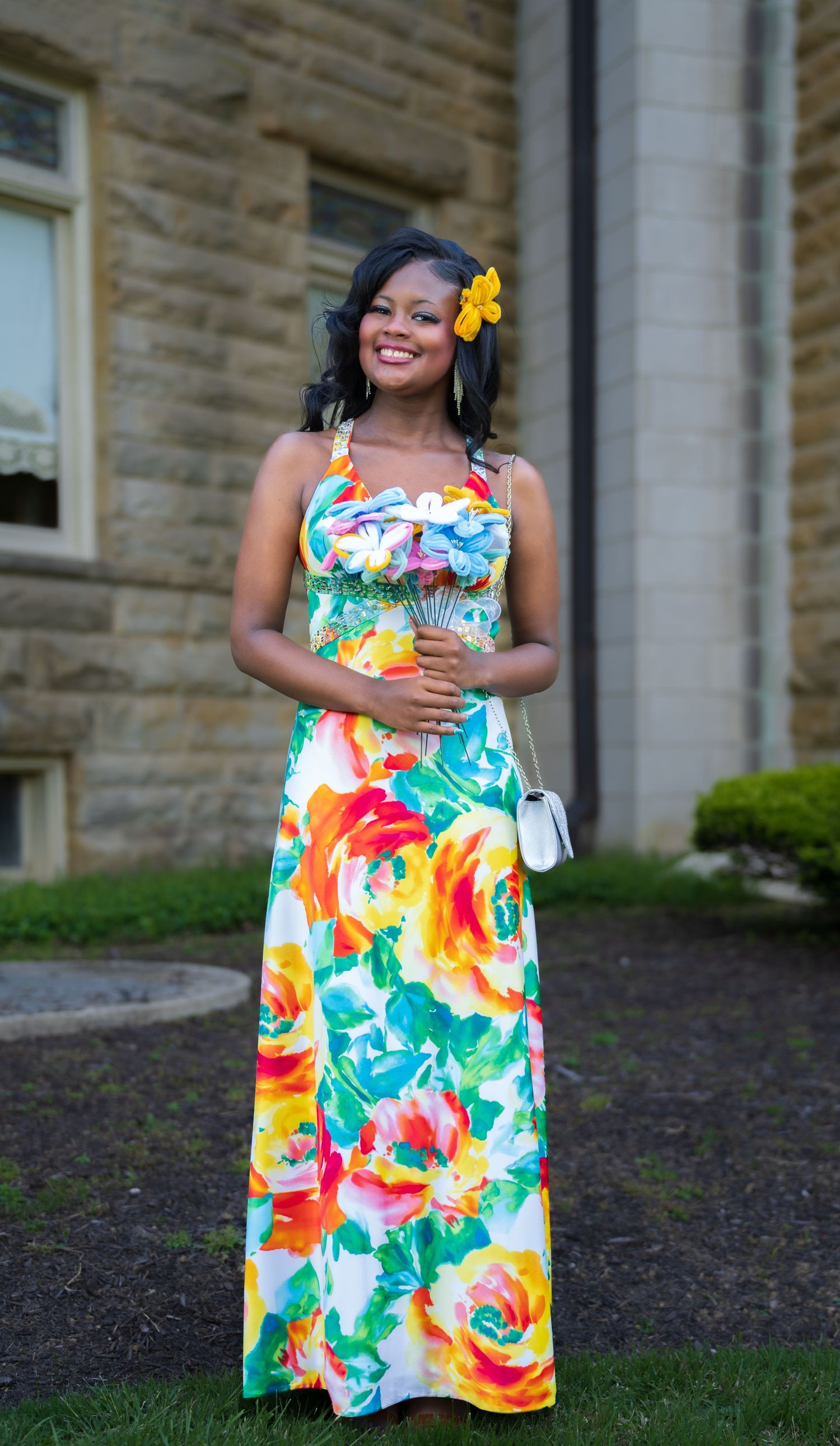 Smiling woman in vibrant floral maxi dress with yellow flower hair accessory outdoors.